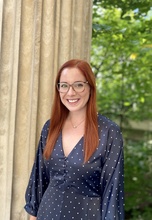 Woman with long red hair, blue dress, and black glasses smiling into the camera with a green and tan background