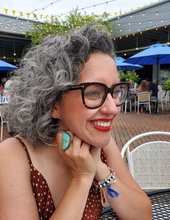 Woman sitting outside with grey hair and red lips with a turquoise ring on her finger. 