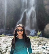 Woman with long flowing hair in teal top standing in front of a waterfall wearing sunglasses.