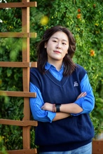 Miao Zhao leaning on a wooden structure, wearing a blue shirt and sweater and looking into the camera